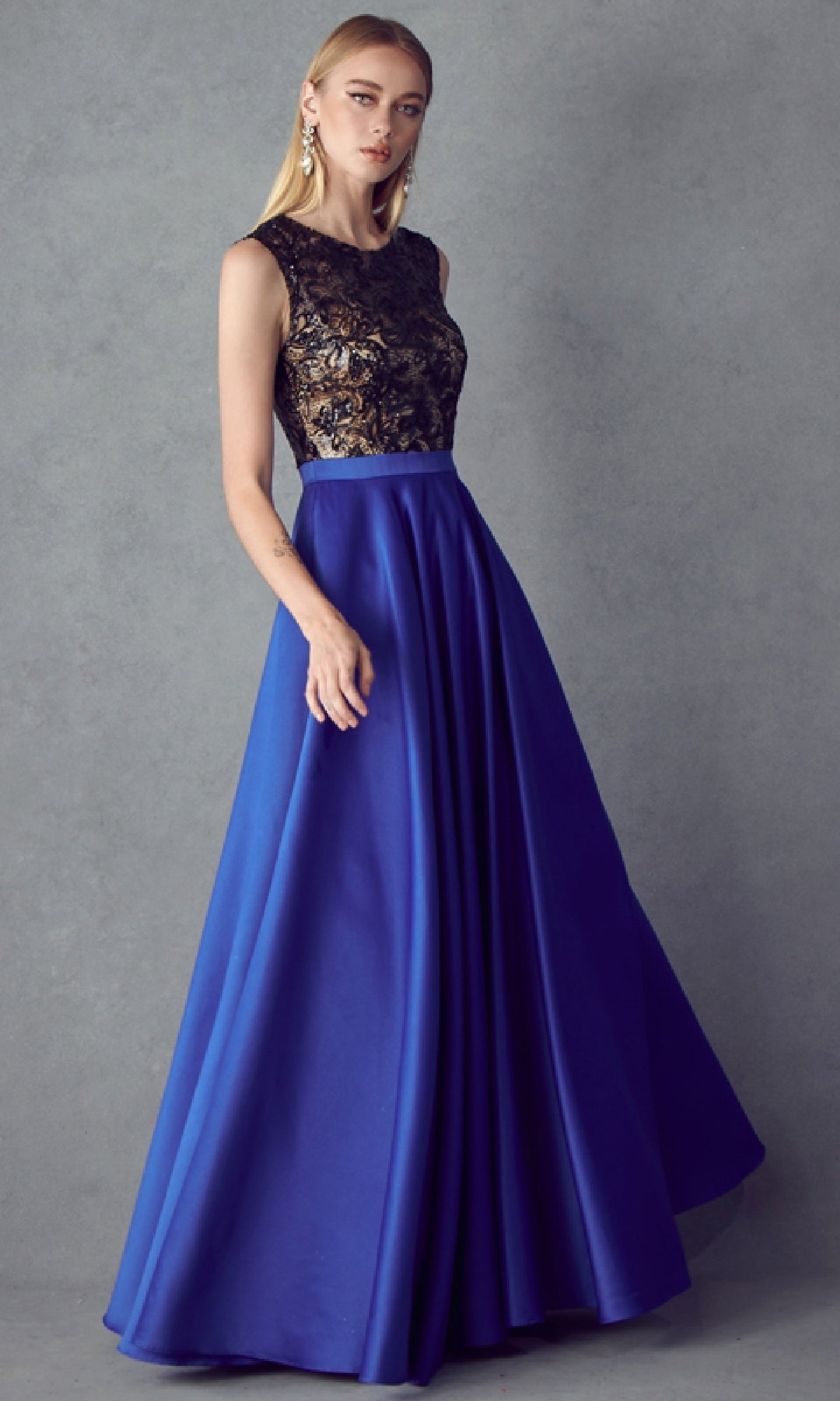 A woman models the Juliet 661 Cut-Out Long A-Line Prom Dress, showcasing a sleeveless black lace bodice and a flowing royal blue skirt against a gray background.